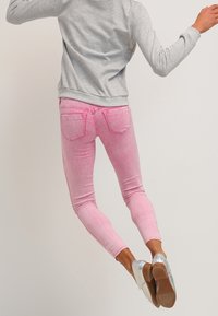 Henkilö hyppäämällä yllään harmaa pitkähihaine paita, vaaleanpunaiset kapeat farkut ja läpinäkyvät korkokengät yksivärisen taustan edessä.