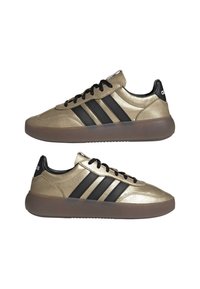 Zapatillas sintéticas doradas con franjas laterales negras, acabado texturizado, cordones negros y una suela translúcida marrón. Cuenta con un collar acolchado.
