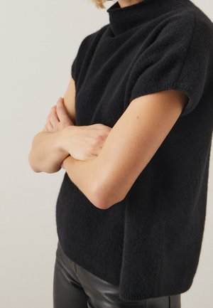 Personne aux bras croisés portant un pull noir texturé à manches courtes et un pantalon en cuir noir, sur un fond uni.