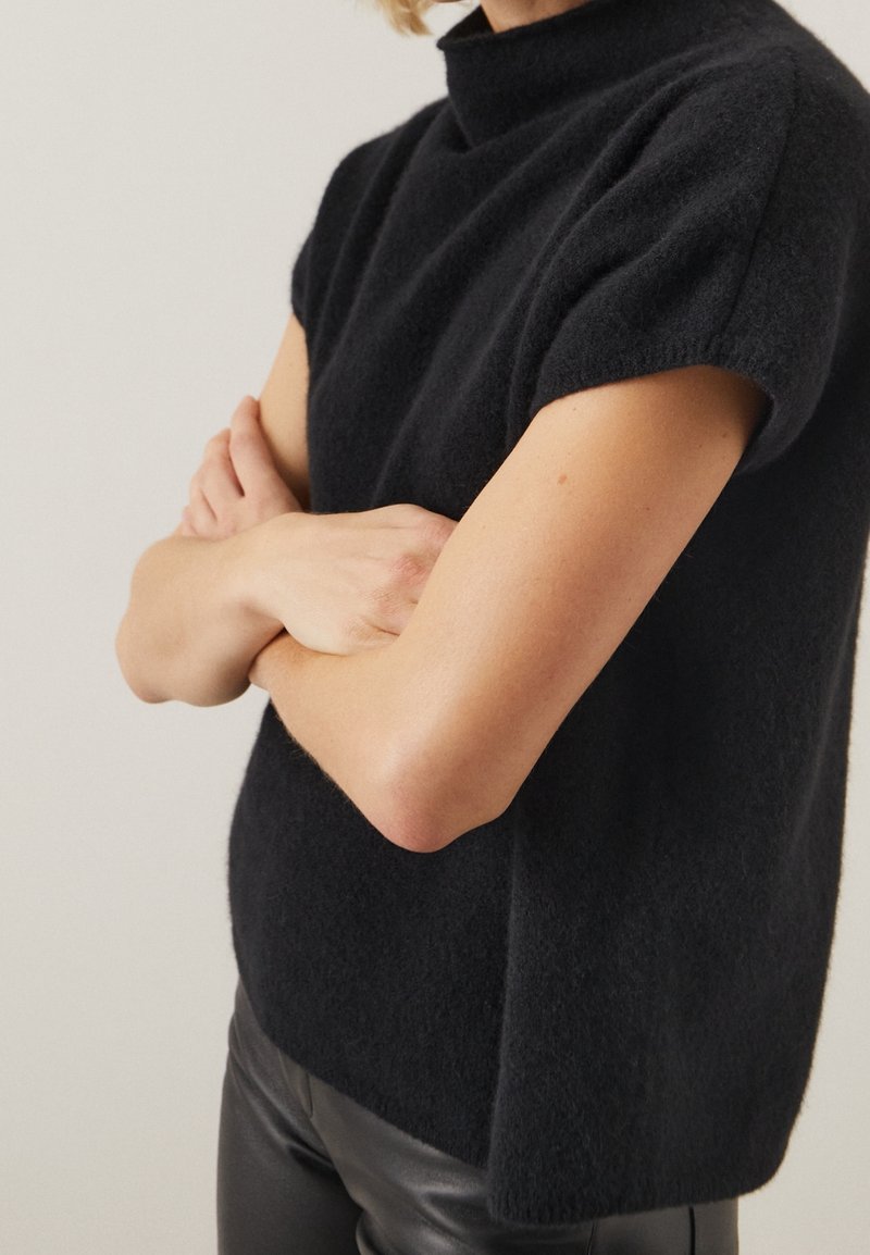 Personne aux bras croisés portant un pull noir texturé à manches courtes et un pantalon en cuir noir, sur un fond uni.