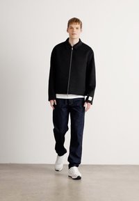 Veste noire zippée avec un col, associée à une chemise blanche à plusieurs couches et un pantalon sombre. Le modèle porte des baskets blanches. Textures lisses.