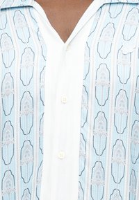 Πουκάμισο με ανοιχτό γαλάζιο και λευκό κάθετο μοτίβο, λευκή ρίγα με κουμπιά, τρία ορατά κουμπιά και λευκό καρφίτσα πεταλούδα στη δεξιά πλευρά.