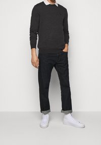 Suéter de punto carbón con borde acanalado, superpuesto a una camisa blanca con cuello. Jeans oscuros de mezclilla, con puños enrollados, complementados con zapatillas blancas.