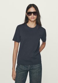 T-shirt in cotone grigio scuro con maniche corte, scollo rotondo, texture liscia e vestibilità rilassata, abbinato a occhiali da sole scuri e jeans blu.