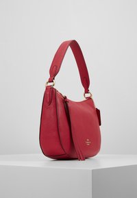 Sac à main en cuir rouge avec une forme incurvée, une quincaillerie dorée et une finition texturée. Il est doté d'une sangle courte et d'un détail à pampille.