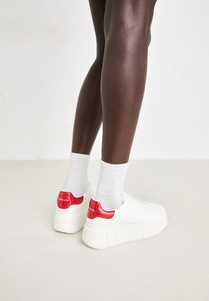 Weiße Sneakers mit einem glänzenden roten Lackleder-Fersenbereich und strukturiertem Sohlen, kombiniert mit weißen gerippten Socken, die ein lässiges Design hervorheben.