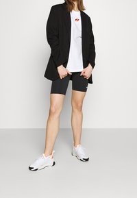 Person som bär svart blazer, vit t-shirt med röd läppavtryck, svarta Nike cykelshorts och vita sneakers med orangea detaljer.