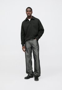 Sudadera negra con cremallera y cuello, combinada con jeans de mezclilla grises. La modelo está de pie con las manos relajadas, usando zapatos negros.