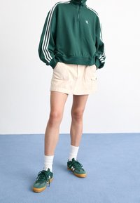 Groene rits-trui met witte strepen, beige shorts met zakken, witte sokken en groene sneakers met rubberen zolen.