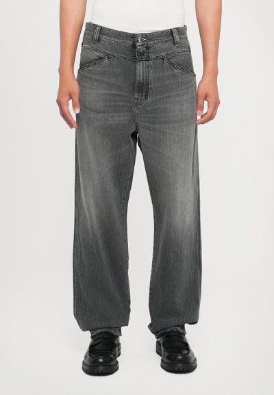 Bredde ben grå denimbukser med en falmet finish. Har fem lommer, bæltestropper og en knaplukning. Båret med sorte sko.