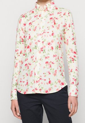 Chemise blanche à manches longues boutonnée avec un motif floral rose et rouge portée avec un pantalon noir sur une personne montrée des épaules aux hanches.