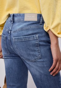 Gros plan d'une personne portant un jean en denim bleu et un pull jaune texturé, mettant l'accent sur la poche arrière et la zone de la hanche.