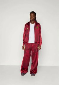 Bordo satino treniruotės kostiumas su auksiniais akcentais, įskaitant užtrauktuku užsisegančią striukę su trimis juostelėmis ir logotipu, derinama su plačiomis kelnėmis.