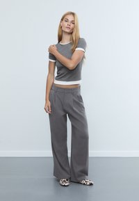 Grauer, taillierter T-Shirt mit weißem Besatz, kombiniert mit grauen, weit geschnittenen Hosen. Das Model trägt gemusterte Slipper mit schwarz-weißen Akzenten.