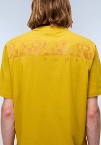 Person with shoulder-length hair wearing a yellow Napapijri T-shirt, shown from the back, against a plain light background.