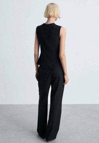 Top sans manches noir, taillé sur mesure, avec une fermeture éclair discrète à l'arrière, associé à un pantalon large noir. Tissu lisse et design structuré.
