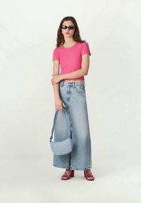 Jovem mulher a usar óculos de sol pretos, uma blusa rosa com riscas e mangas curtas, uma saia comprida de ganga clara, sandálias cor de rosa e a segurar uma mala azul clara.