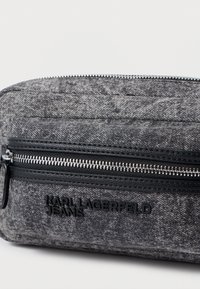 Detailný záber na šedý textúrovaný látkový sáčok s čiernym zipsom a logom "Karl Lagerfeld Jeans" v čiernych písmenách na prednej vrecku.