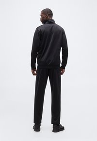 Άνδρας που στέκεται γυρισμένος με την πλάτη φορώντας μαύρο αθλητικό μπουφάν, ταιριαστό παντελόνι και μαύρα αθλητικά παπούτσια σε λευκό φόντο.