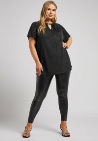 Mustapaitainen lyhythihainen toppi, jossa on sormivaihtoehto kaula-aukossa ja hillitty glitter yksityiskohta, yhdistettynä kiiltäviin mustiin legginsseihin ja remmikorkokengiin.
