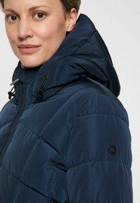 Nahaufnahme einer Person, die eine marineblaue gepolsterte Jacke mit Kapuze und einem runden schwarzen Logo auf dem oberen Ärmel trägt.