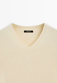 V-hals trui in neutrale kleur, gemaakt van een zachte breimateriaal. Beschikt over ribgebreide randen en een klein logo-label aan de binnenkant van de kraag.