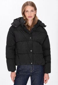 Sort pufferjakke med cropped design, trykknapper, justerbar hætte, quiltet tekstur og lange ærmer, båret med mørke jeans.