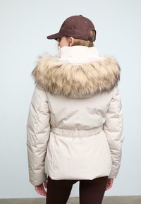 Giacca puffer color crema con colletto in pelliccia sintetica, vita aderente e dettagli elasticizzati; abbinata a un cappello marrone.