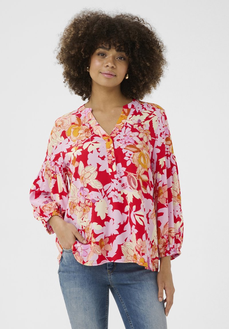 Gėlių bluzonas su raudonu ir rožiniu raštu, turintis ilgas pūkuotas rankoves ir V formos iškirptę. Pagamintas iš lengvos medžiagos, siūlantis laisvą siluetą.