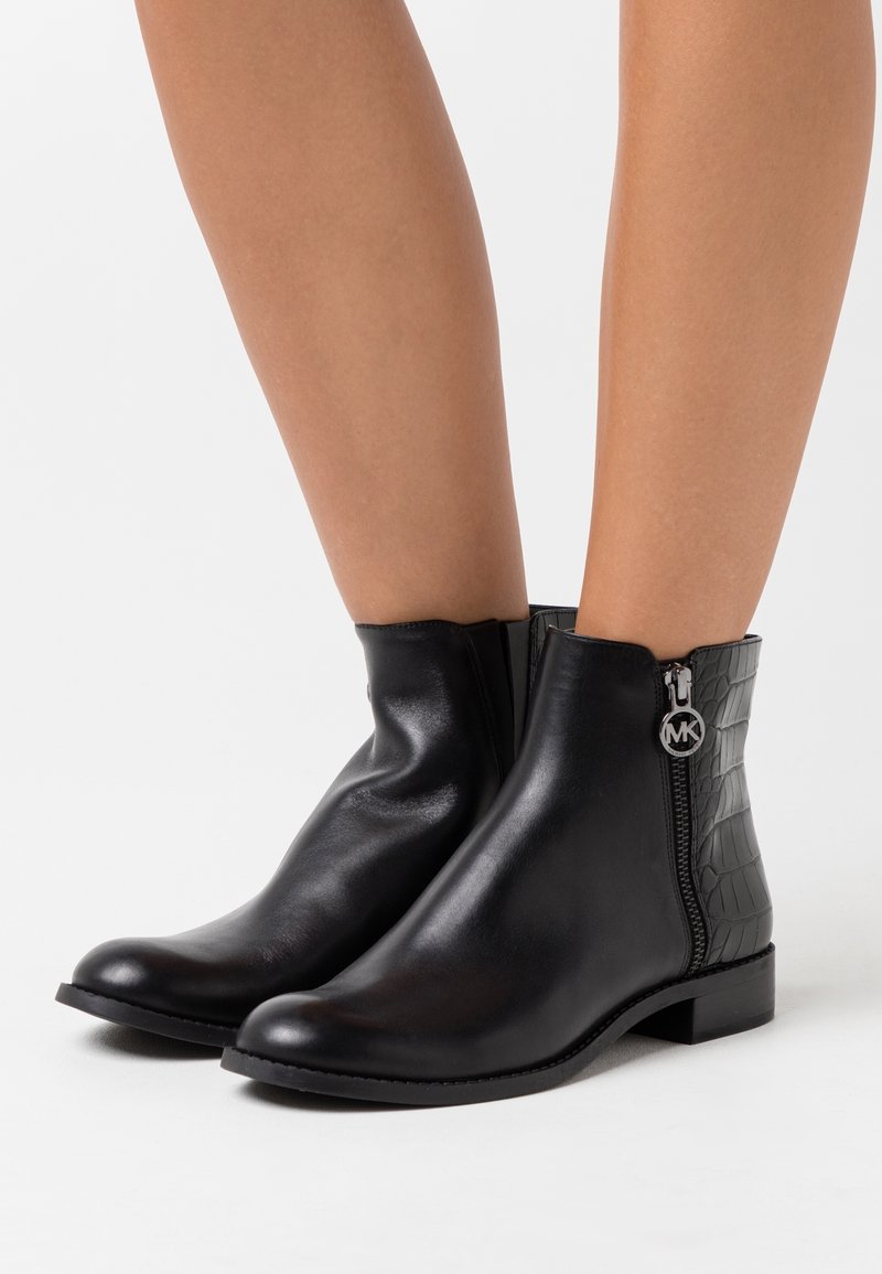 Bottes à cheville en cuir noir avec fermeture éclair latérale, ornées d'un pendentif rond en argent avec le logo MK, portées sur des jambes nues sur un fond blanc.