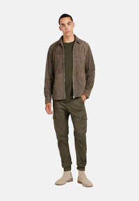 Giacca in suede marrone con zip, camicia verde oliva, pantaloni cargo verde oliva con polsini elastici e stivali alla caviglia beige chiaro. Dettagli di design semplici.