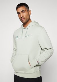 Light green hoodie made of cotton blend fabric, featuring a front kangaroo pocket and embroidered "Champion" logo on the chest.