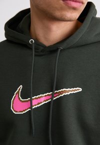 Mørkegrøn hættetrøje med et stort, pixeleret Nike swoosh i pink og brun. Stof ser blødt ud med justerbare snore og metaløjer.