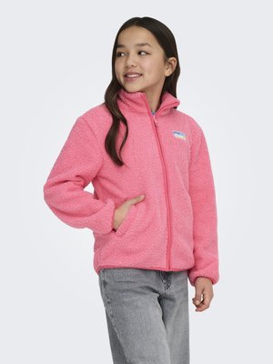 Ung jente iført en knallrosa fleecejakke med frontlommer og glidelås, stående med en hånd i lommen og ser sidelengs.