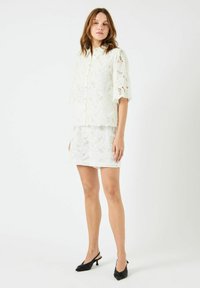 Hvit bluse i blonde med dekorative mønstre, korte ermer og knappestolpe foran, kombinert med et matchende miniskjørt i blonde og svarte hæler.