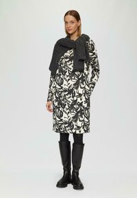 Robe à manches longues avec un motif floral noir sur un tissu blanc, assortie à une écharpe en tricot gris foncé et des bottes noires hautes.
