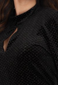 Czarna aksamitna odzież z wysokim kołnierzem, z diamentowym wzorem z wielobarwnymi błyskotkami. Widoczny detal w postaci dekoltu w kształcie dziurki.