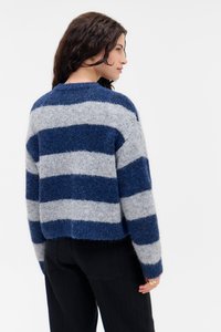 Vrouw in een fuzzy blauw-grijs gestreept sweater en zwarte broek, van achteren getoond en iets naar de zijkant gedraaid.