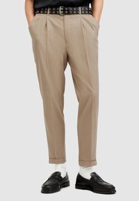 Pantaloni beige con un design plissettato, abbinati a una cintura nera con fori circolari e mocassini neri. Calzini bianchi sono visibili.