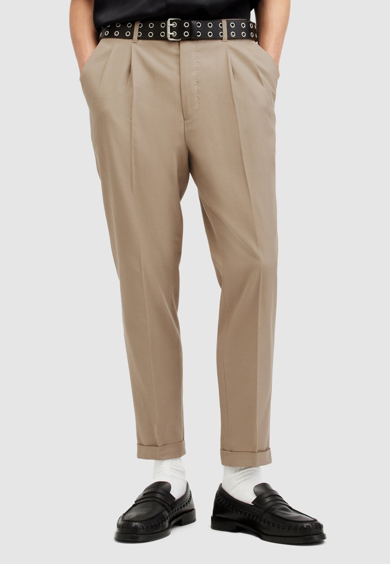 Pantaloni beige con un design plissettato, abbinati a una cintura nera con fori circolari e mocassini neri. Calzini bianchi sono visibili.
