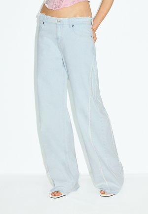 Modèle portant un jean bleu clair à jambes larges avec des ourlets bruts et des coutures latérales, associé à des talons blancs ouverts à l'avant et un top court rose.