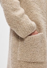 Manteau en beige texturé avec un tissu pelucheux, une coupe décontractée et une poche latérale avec un détail cousu sur le devant.