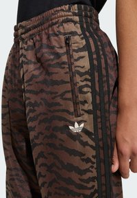 Smeđe i crne trenirka hlače s uzorkom tigrovih pruga, elastičnim pojasom, bočnim džepom na patentni zatvarač i bijelim logotipom na bedrima; tkanina s teksturom.