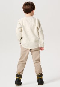 Cremefarbener Pullover mit gesprenkeltem Muster, kombiniert mit beigen Hosen. Das Kind trägt schwarze Stiefel mit gelben Akzenten. Das Bild zeigt die Rückansicht.