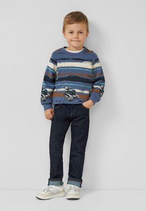 Jonge jongen staand, gekleed in een blauw gestreepte trui met voertuigen, donkere spijkerbroek en witte sneakers, tegen een effen achtergrond.