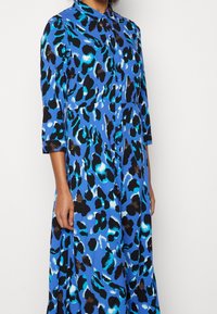 Blaues Kleid mit Leopardenmuster, dreiviertel langen Ärmeln und durchgehender Knopfleiste, aus glattem Stoff mit schwarzen und türkisen Akzenten.