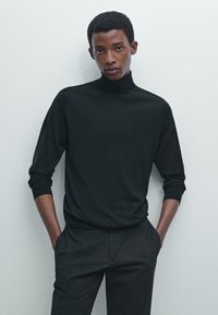 Black turtleneck sweater made of soft fabric with ribbed cuffs and hem, paired with black trousers, against a light background.