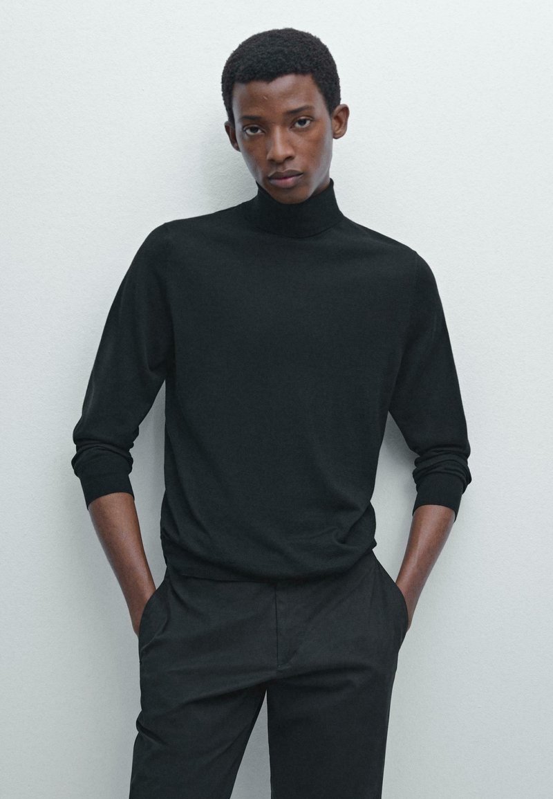 Schwarzer Rollkragenpullover aus weichem Stoff mit gerippten Bündchen und Saum, kombiniert mit schwarzen Hosen, vor einem hellen Hintergrund.