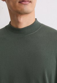 Dunkelgrüner Pullover mit hohem, geripptem Kragen und glatter Textur, der ein tailliertes Design ohne Muster oder Metallakzente präsentiert.