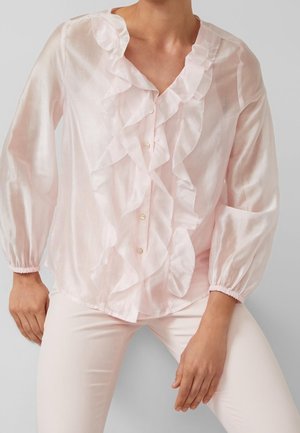 Femme portant un chemisier rose clair transparent avec des volants sur le devant et des poignets élastiques, associé à un pantalon rose clair.